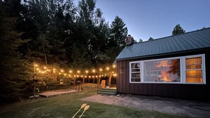 Een chalet met een metalen dak, een terras en lichtjes. Twee houten peddels liggen op het gras.