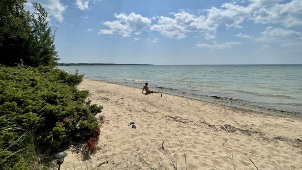 Een strand met iemand die op het zand zit, omgeven door begroeiing en de zee.