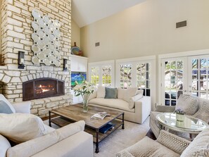 A living room with a stone fireplace, a white sofa, a coffee table, and large windows with a view of the outdoors. - Above Auth Westlake I Pool I Hot Tub I Game Room I Back House I Greenbelt (West Lake Hills)