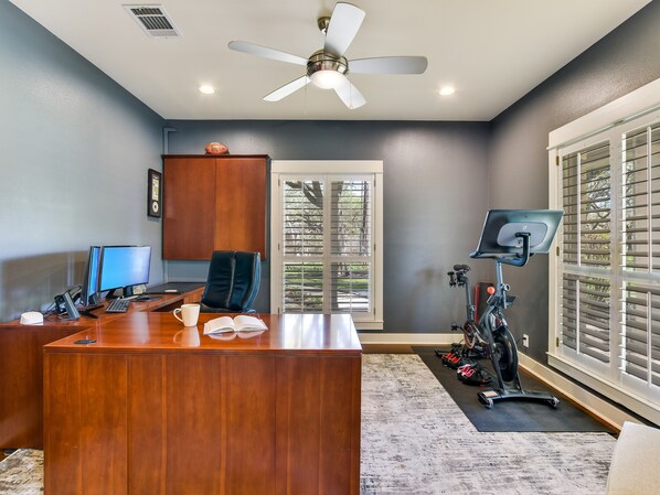 Een werkplek aan huis met een houten bureau, twee computermonitoren, een stoel, een plafondventilator en een fiets.