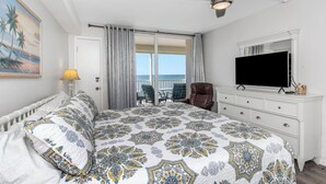 Une chambre avec un lit, un ventilateur de plafond, une télévision, une chaise, une commode et un balcon avec vue sur l’océan.