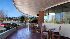 An outdoor patio with wooden tables and chairs, a curved roof, and a view of a body of water and hills in the background. - Hotel Costanera (Villa Carlos Paz)