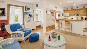 A spacious living room with a wicker sofa, a round glass coffee table, and a kitchen area with wooden cabinets and a white countertop.