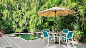 A poolside area with a table and chairs, surrounded by lush greenery.