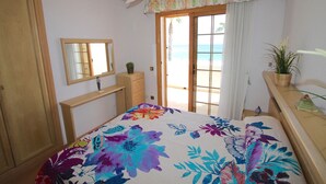 Une chambre avec un lit, une commode, un miroir, un ventilateur de plafond, une fenêtre donnant sur la plage et une plante en pot.