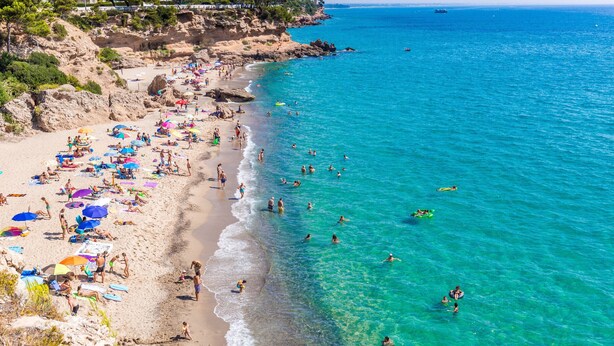 Une plage avec une eau turquoise et limpide, des gens qui se baignent et qui prennent des bains de soleil, et des parasols sur le sable.