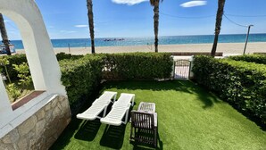 Une terrasse avec une pelouse artificielle, deux chaises longues, une petite table et un portail donnant accès à la plage.