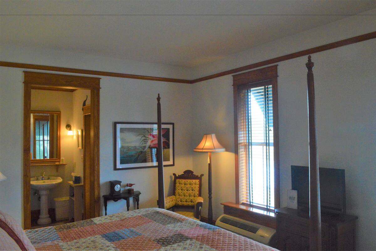Un dormitorio con cama, mesita de noche, silla, lámpara, espejo, ventana con persianas y televisor.