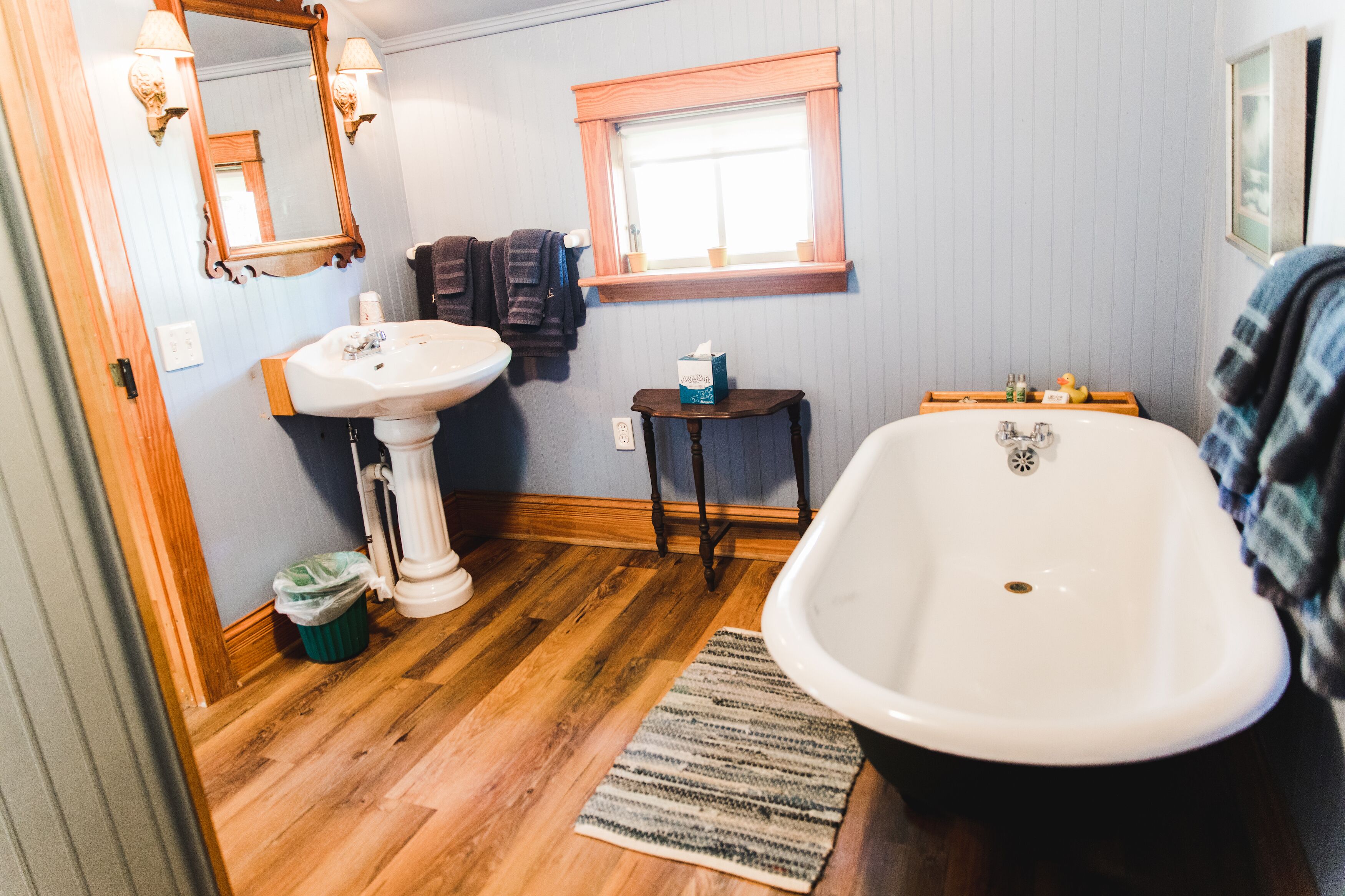 A bathroom with a wooden floor, a white sink, a bathtub, a window with a wooden frame, and a mirror with wall sconces.