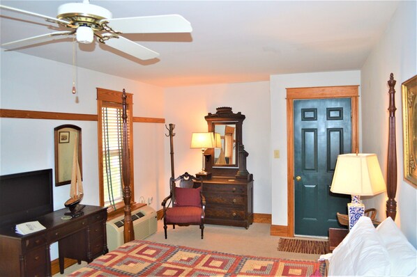 A hotel room with a bed, a dresser, a chair, a desk, a ceiling fan, a door, and a window with blinds. - Roanoke Island Inn (Manteo)
