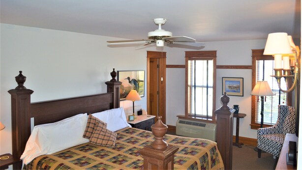 A hotel room with a bed, a ceiling fan, a chair, a table, and a window with blinds. - Roanoke Island Inn (Manteo)