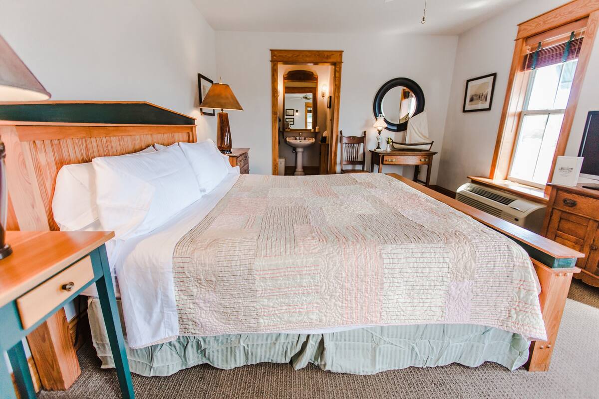 Un dormitorio ordenado con una cama grande, una mesita de noche de madera, un escritorio, una silla, un espejo y una ventana con persianas.
