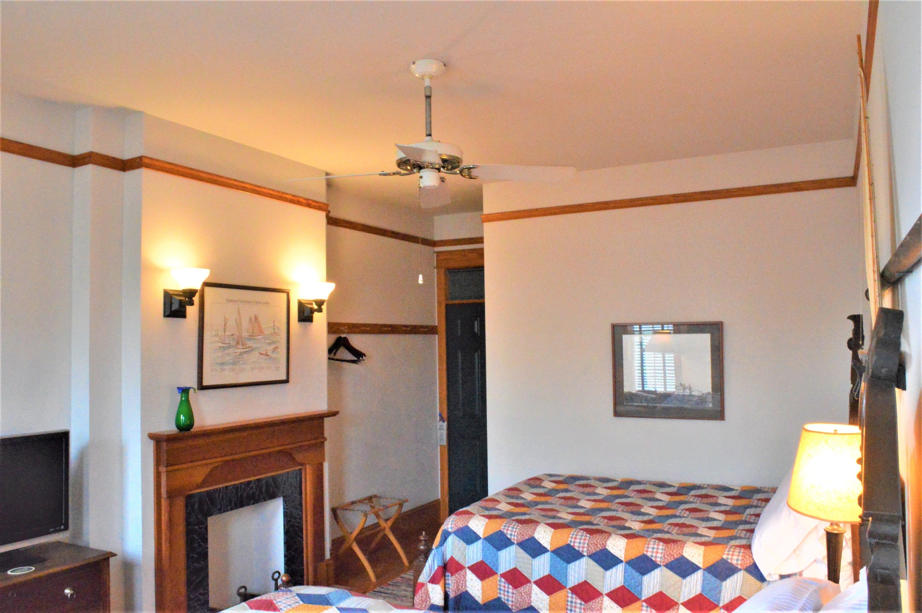 A hotel room with a bed, a fireplace, a ceiling fan, a framed picture, and a window.