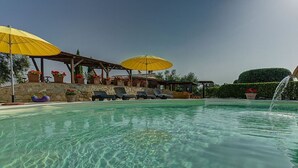Piscina límpida com espreguiçadeiras e guarda-sóis amarelos, rodeada por muros de pedra e vasos de plantas.