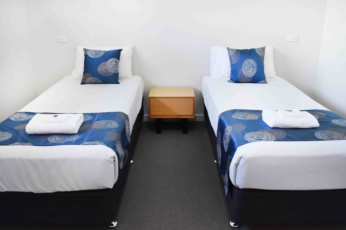 Ein Hotelzimmer mit zwei Einzelbetten, jeweils mit einer blau-weißen Bettdecke und passenden Kissen. Zwischen den Betten steht ein Nachttisch, und auf jedem Bett liegt ein gefaltetes Handtuch.