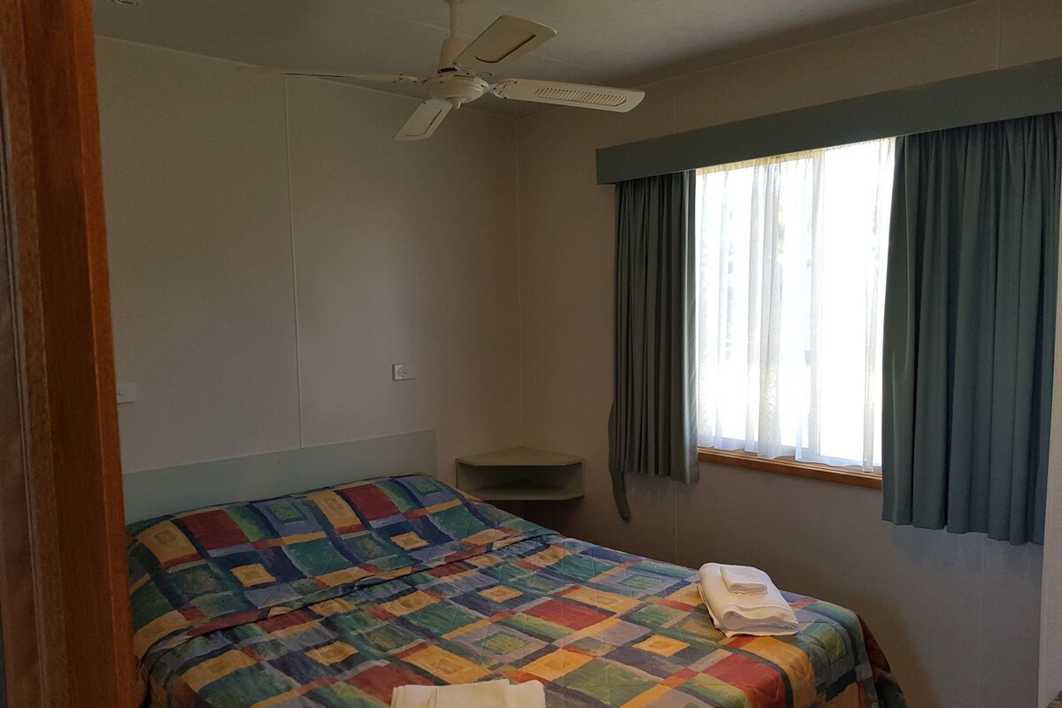 Ein Schlafzimmer mit einem Bett, einem Deckenventilator, einem Fenster mit Vorhängen und einer kleinen Ablagefläche.