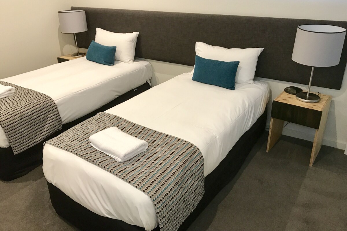 Ein Hotelzimmer mit zwei Einzelbetten, jeweils mit weißer Daunendecke und gemustertem Überwurf. Zwei Nachttische mit Lampen und ein kleiner Holz-Nachttisch mit Lampe sorgen für Licht.