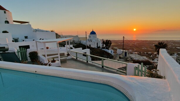 Vue depuis la piscine sur un bâtiment blanc surmonté d’un dôme bleu, qui surplombe un paysage côtier au coucher du soleil.