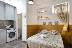 Une chambre d’hôtel compacte comprenant un lit, une table de chevet, une petite commode, un lave-linge et une petite kitchenette.