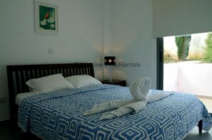 Ein Schlafzimmer mit einem Bett, einem Nachttisch, einer Wandleuchte und einem Fenster mit Blick auf Grünflächen.