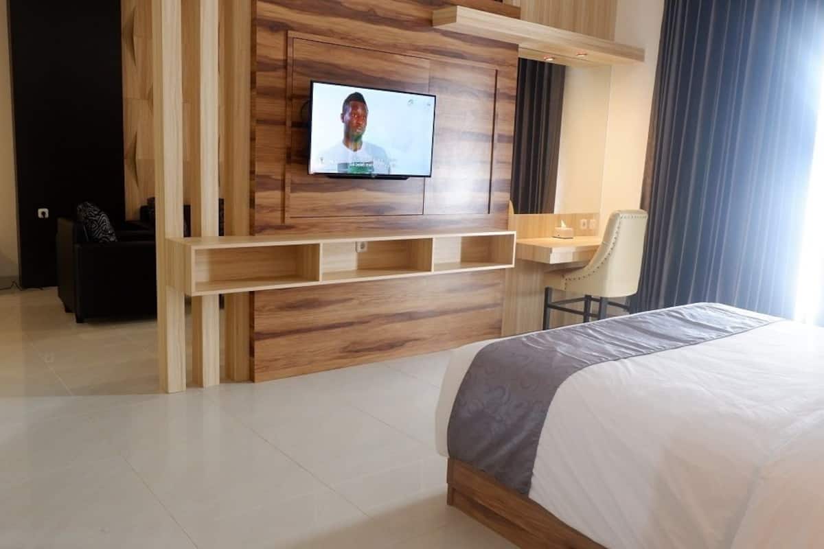 A modern hotel room with a large bed, a TV mounted on a wooden wall, a desk with a chair, and a window with curtains.