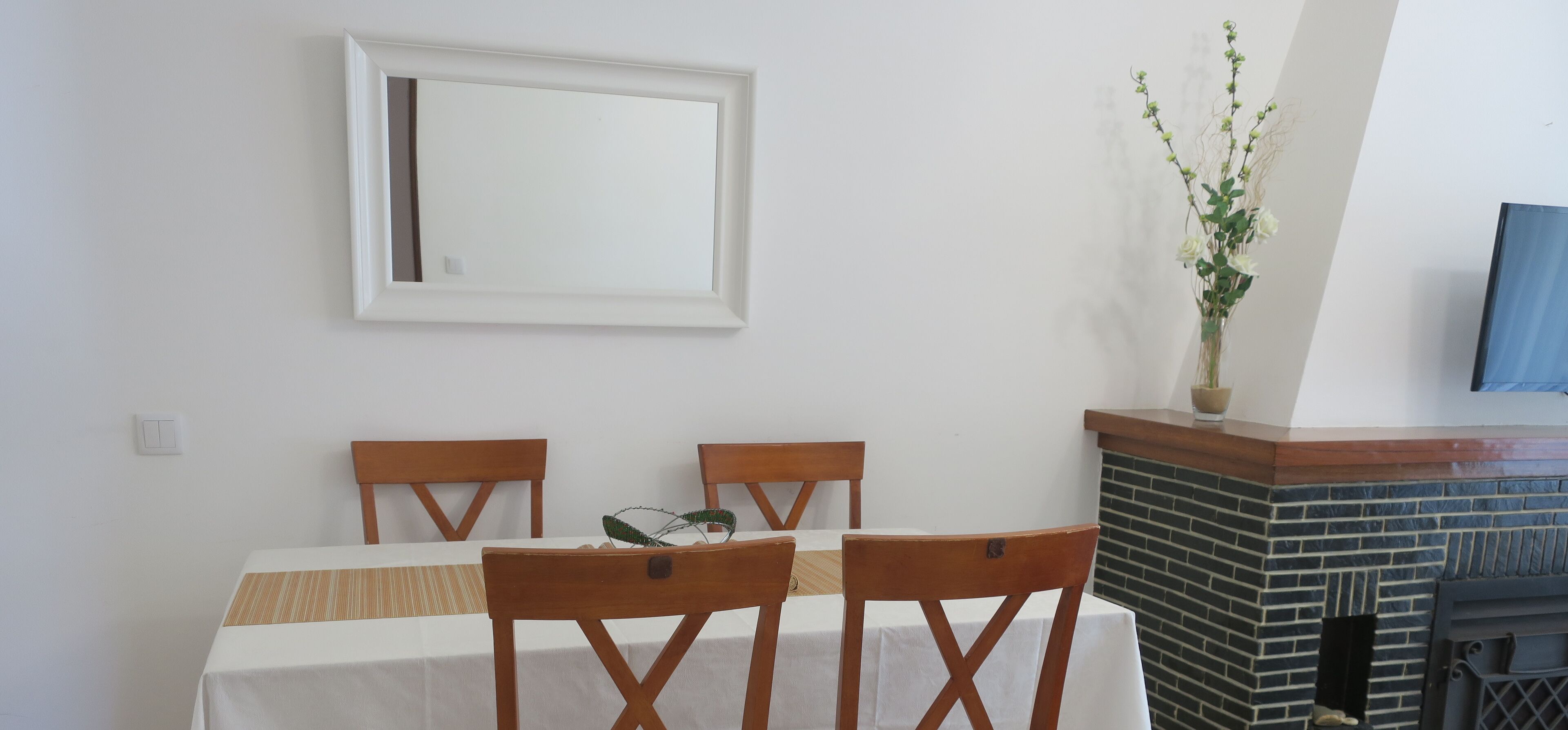 A dining area with a table set for three, wooden chairs, a white tablecloth, a vase with flowers, and a brick fireplace.