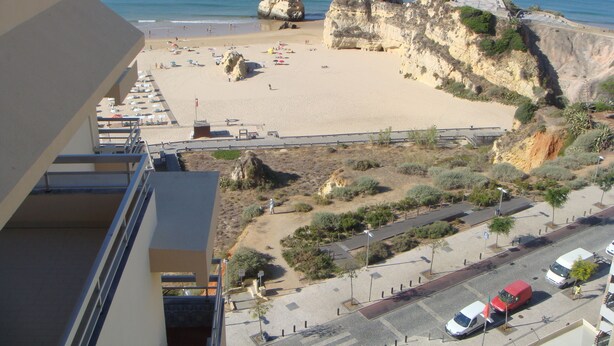 A beach with rocky formations, a parking area, and a road with vehicles. -  Wonderful view of the beach and river,  front line with garage T2. 1978 / AL (Praia da Rocha Portimão)