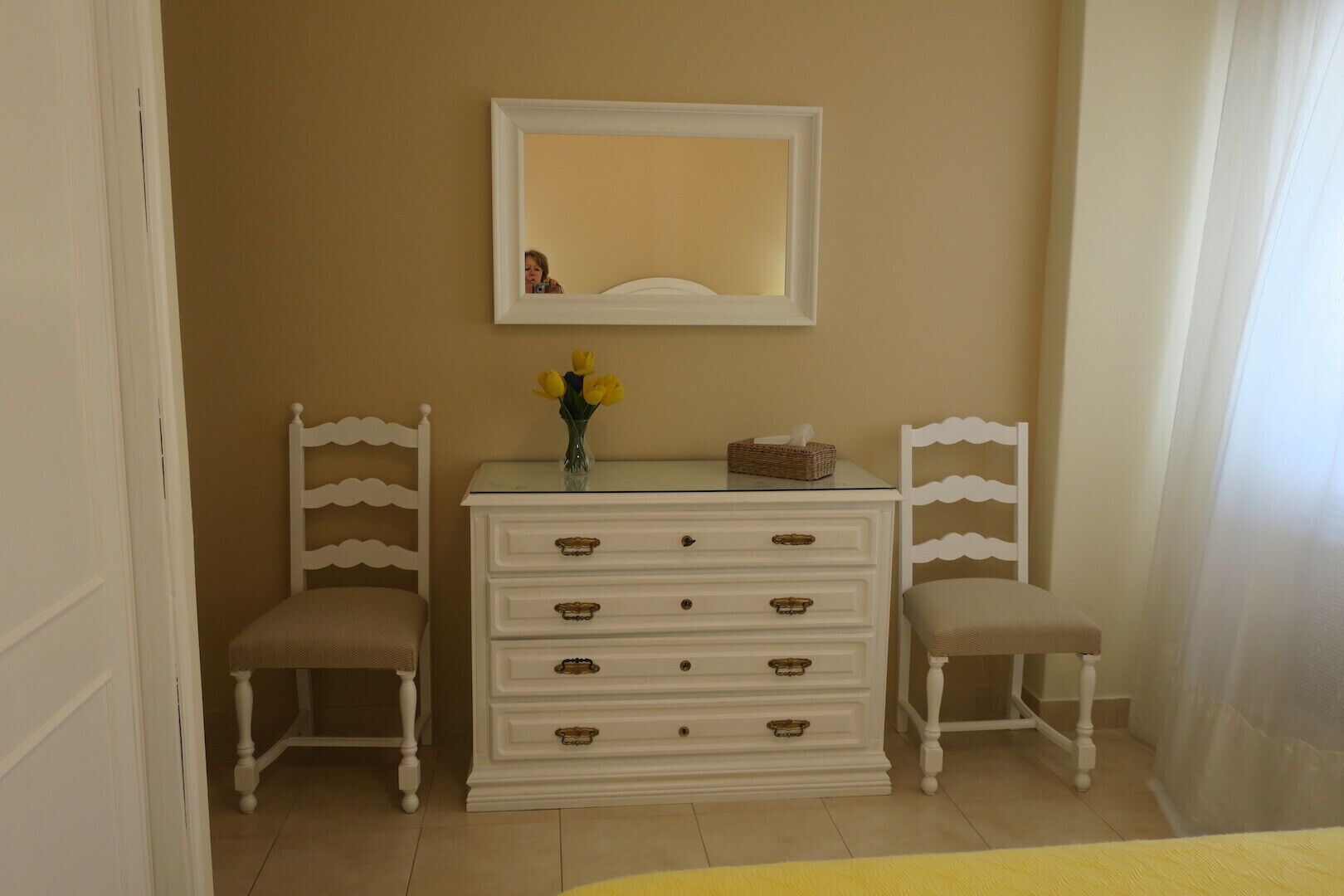A room with a white dresser, two chairs, a mirror, and a vase with flowers.