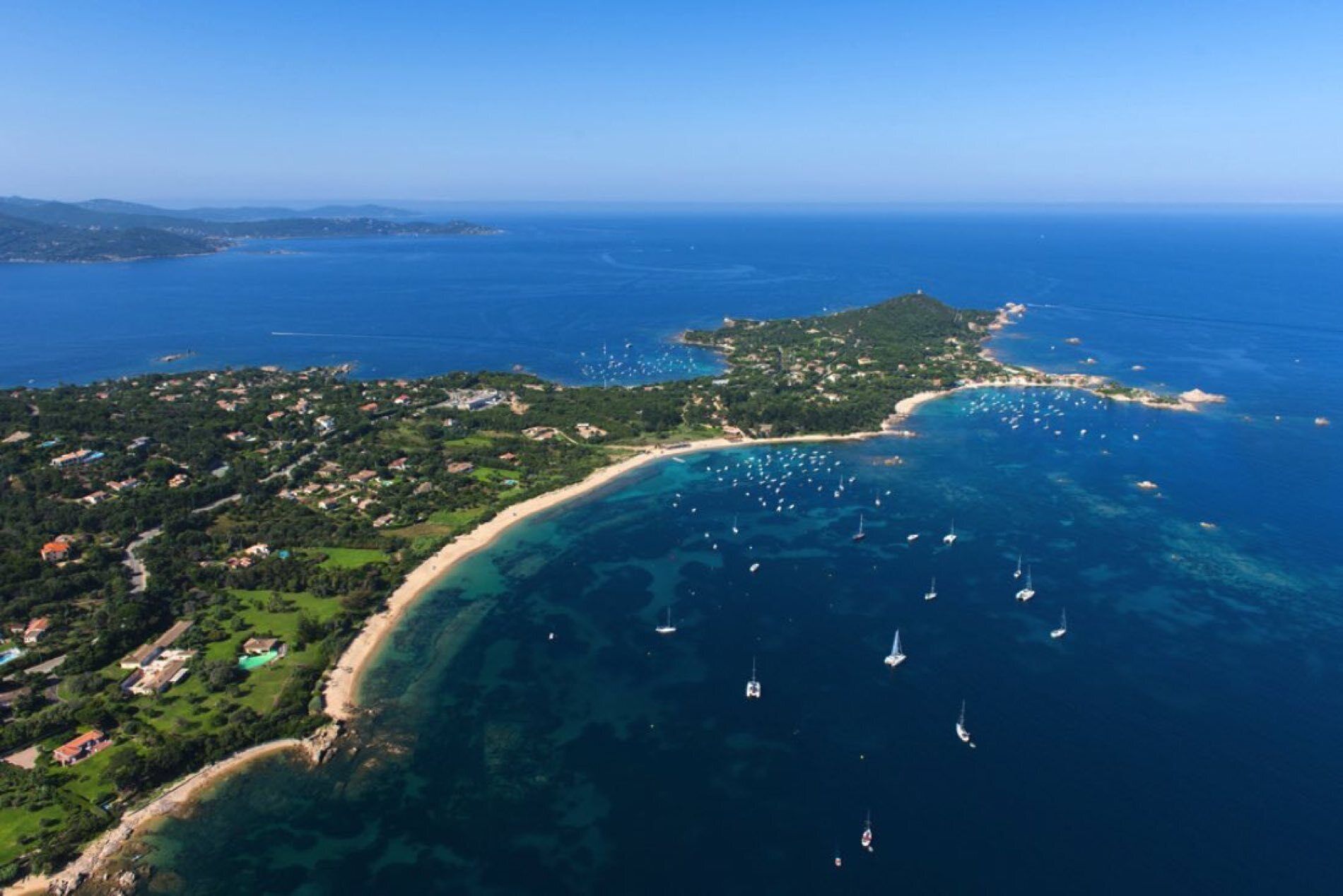 An aerial view of a coastal area with a clear beach, boats in the water, and a hilly landscape with scattered buildings.