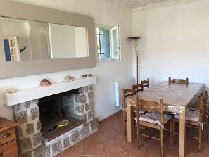 A room with a stone fireplace, a dining table with chairs, a mirror above the fireplace, and a window with a view of the outside.