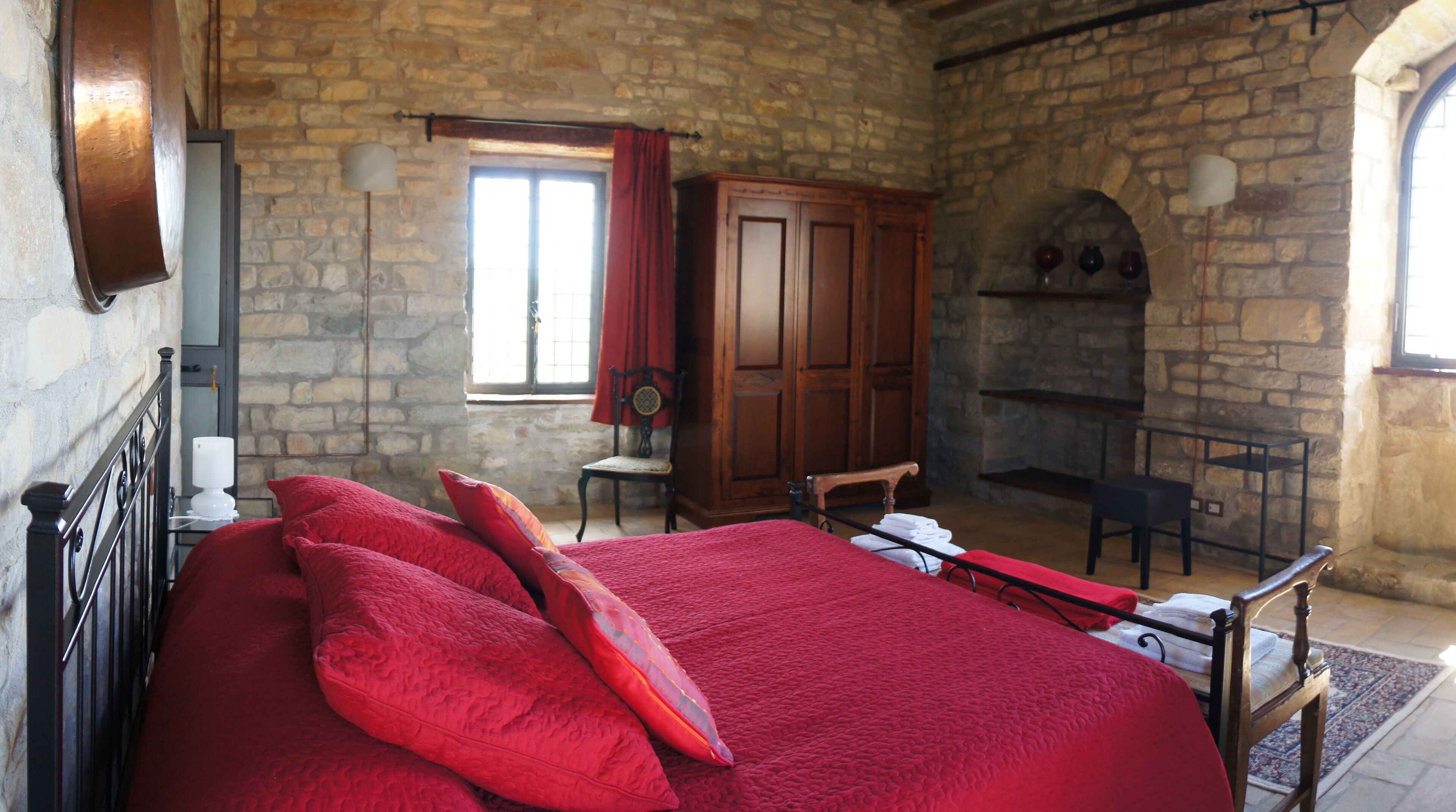 Une chambre avec un mur en pierre, une armoire en bois, une cheminée et un lit avec une literie rouge.