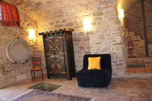 Une pièce aux murs de pierre, dotée d’une cheminée rustique, d’un meuble en bois, d’une chaise et d’un canapé agrémenté d’un coussin jaune.