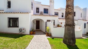 Un edificio blanco con un sendero de adoquines que conduce a una puerta, rodeado de macetas con plantas y una palmera.