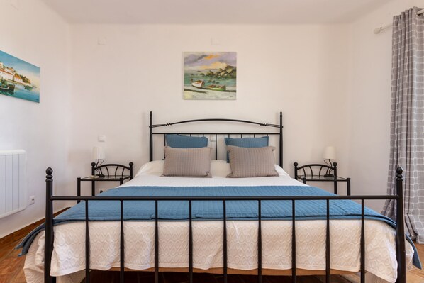 A bedroom with a bed, two bedside tables, a nightstand, a painting on the wall, and a window with curtains. - Casa Borboleta - Communal pool, sea views & 350m from beach & town (Carvoeiro)