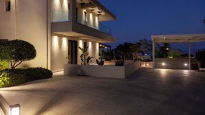 A modern multi-story building with a flat roof, large windows, and a well-lit entrance area with potted plants and a covered seating area. - Orama Luxury Villa, 4 Bedrooms with Private Pool, Sleeps 8 (Piskopiano)
