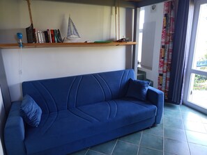 A blue sofa with a cushion, a shelf with books and a model sailboat, and a view through a window with curtains.
