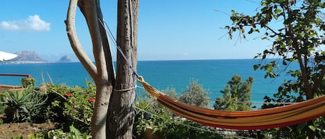 A hammock tied to a tree, overlooking a coastal view with a clear sky and distant mountains.