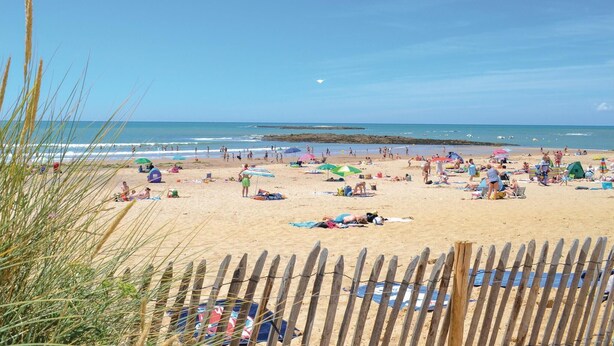 Uma praia com pessoas tomando sol e nadando, uma cerca de madeira em primeiro plano e o oceano ao fundo.