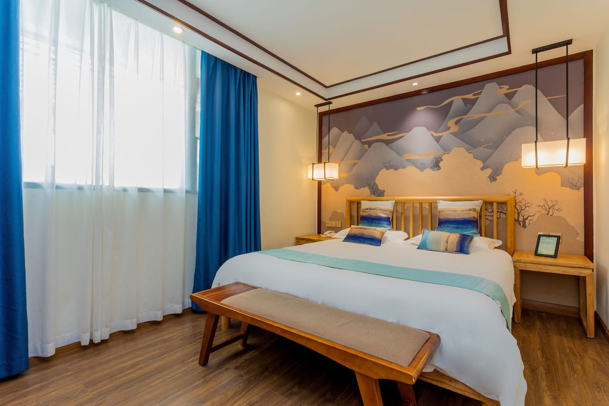 A hotel room with a large bed, wooden headboard, bedside tables, a bench, and a wall mural.