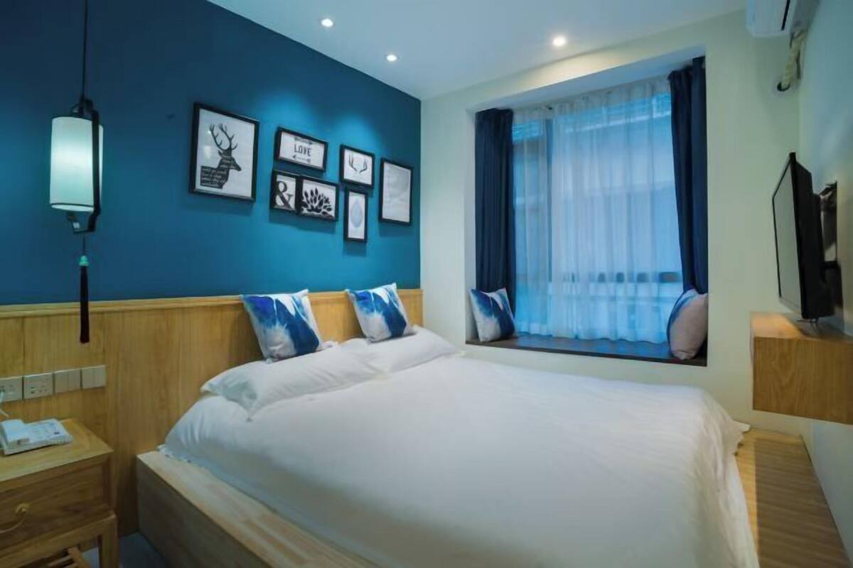 A hotel room with a bed, bedside tables, a window with curtains, wall art, and a ceiling light.