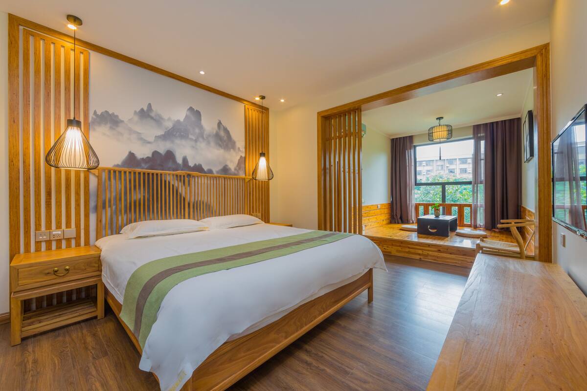 A modern hotel room with a large bed, wooden flooring, a painting of mountains, and a view of greenery through the window.