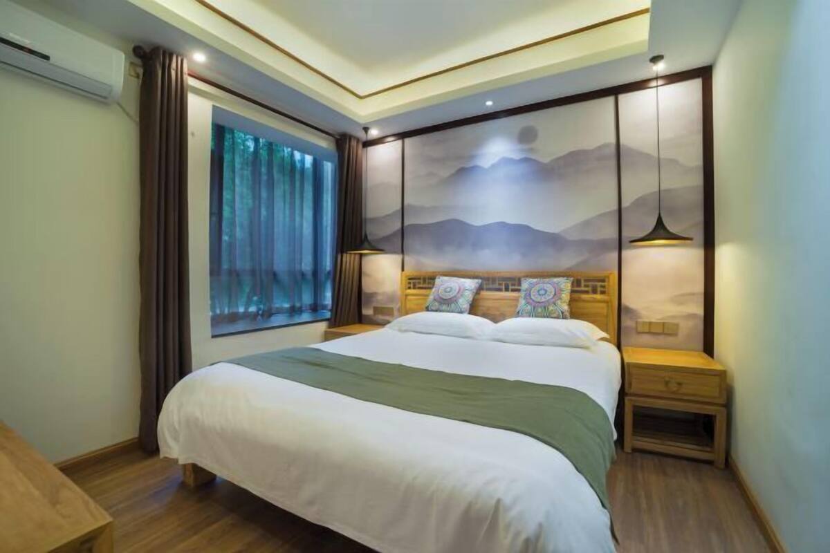 A bedroom with a large bed, a window with curtains, a wall-mounted artwork, and two bedside lamps.