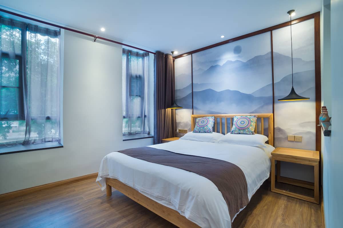 A bedroom with a large bed, a view of the outdoors through a window, and a wall with a mountain painting.