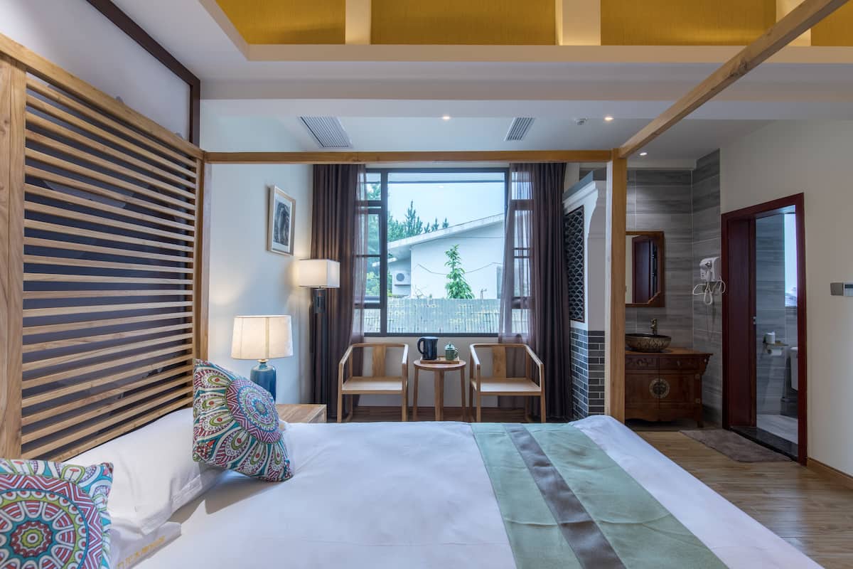 A hotel room with a bed, two chairs, a small table, a lamp, and a view of the outside.