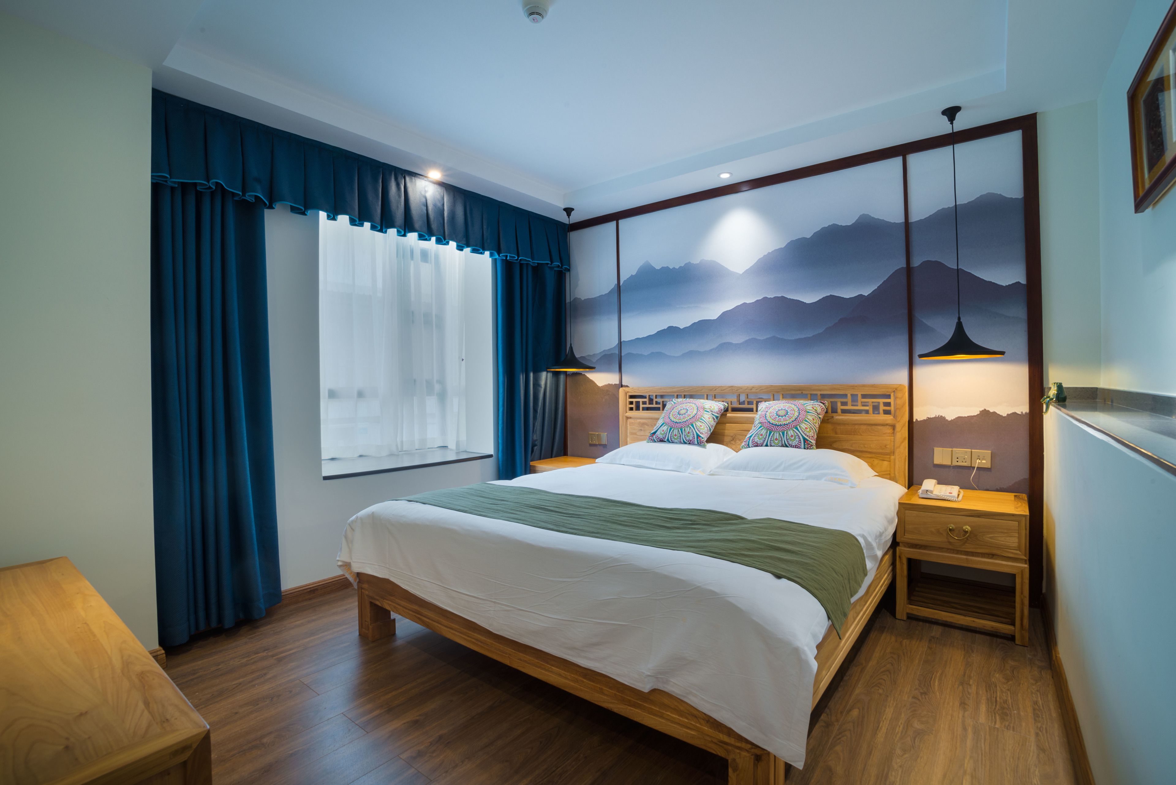 A bedroom with a large bed, a nightstand, a wooden headboard, a window with curtains, and a wall-mounted artwork of mountains.