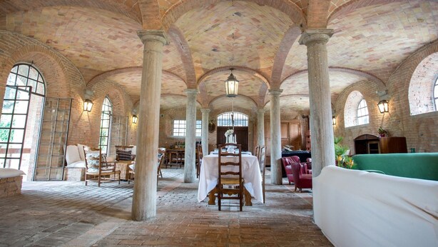 Un intérieur spacieux avec des plafonds en voûte et des murs en briques, comprenant un coin repas central avec une table et des chaises, ainsi que de grandes fenêtres laissant entrer la lumière naturelle.