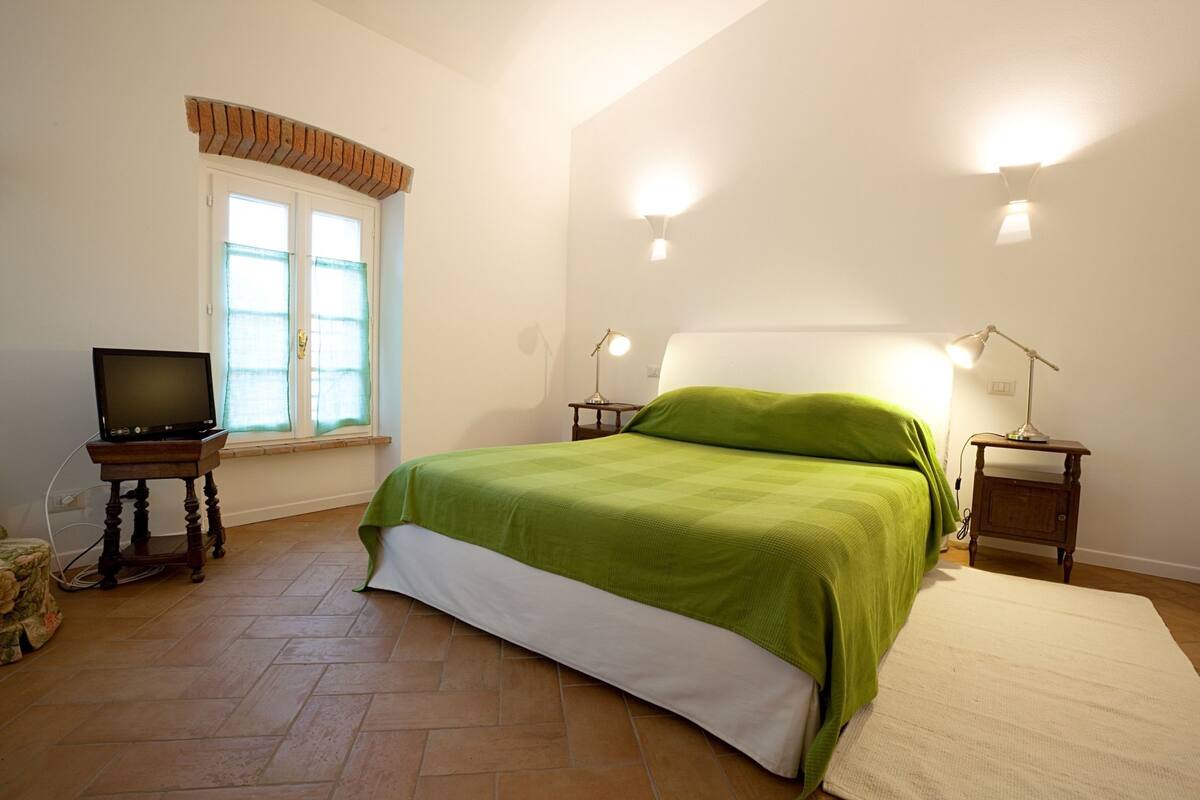 A bedroom with a bed, a television on a stand, a window with curtains, and two bedside lamps.
