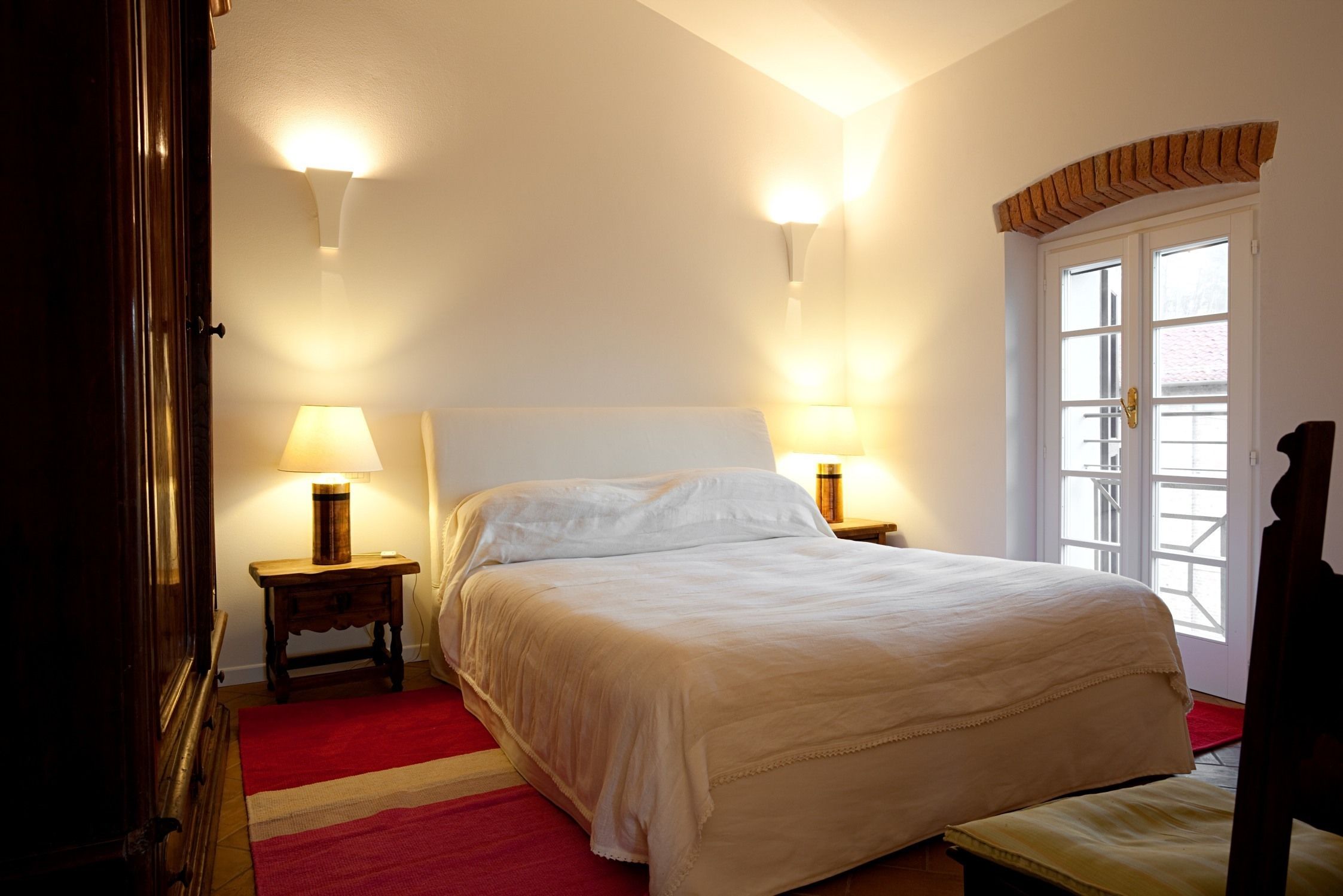 Une chambre avec un grand lit, deux lampes de chevet, une moquette rouge et une fenêtre surmontée d’une arche en briques.