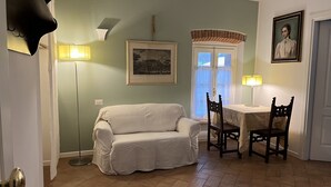 Une pièce avec un canapé blanc, une table à manger avec des chaises, un lampadaire, un tableau encadré au mur et une fenêtre donnant sur un paysage.