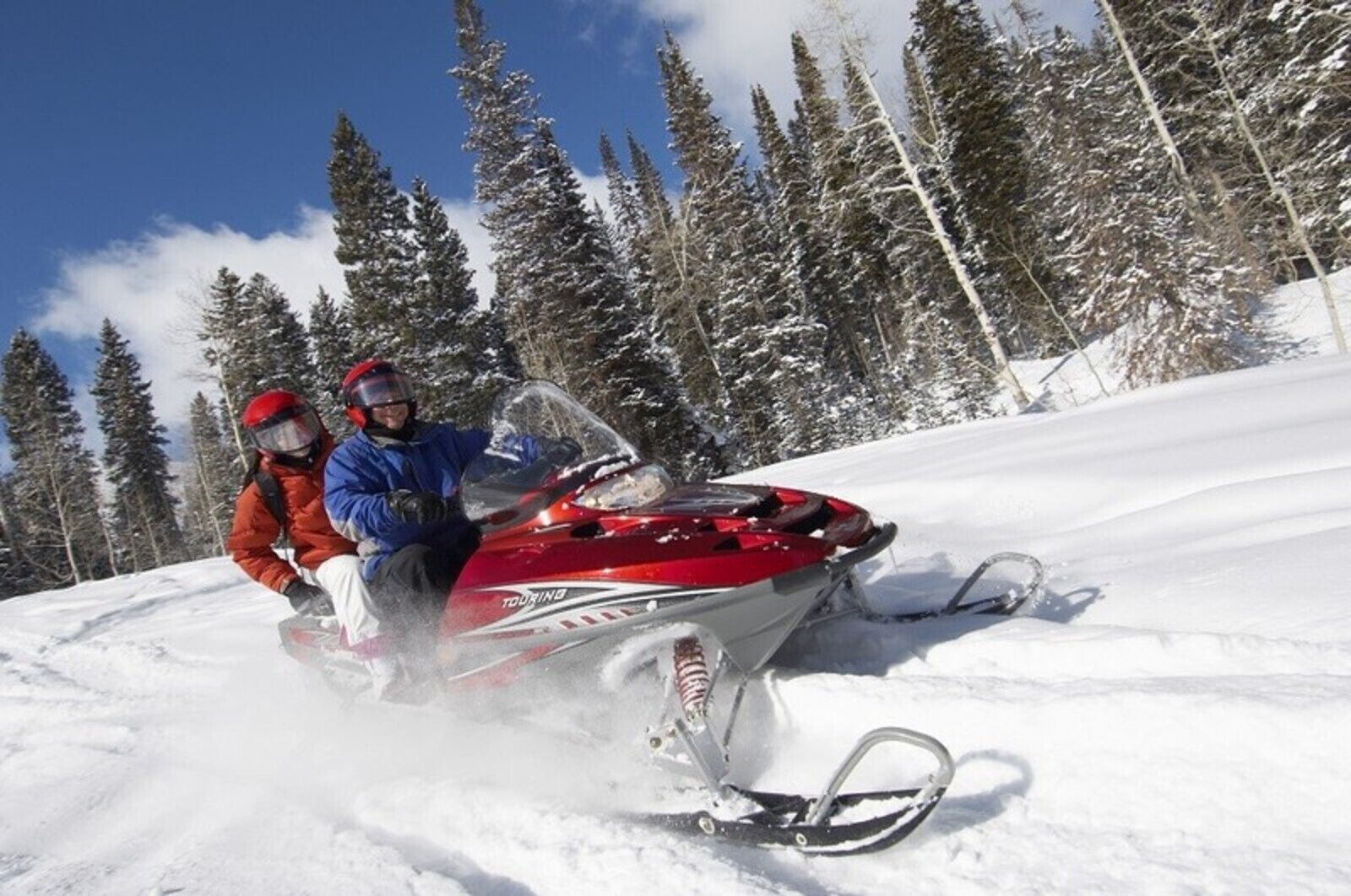 Motocicleta de nieve con dos pasajeros, rodeada de nieve y árboles.